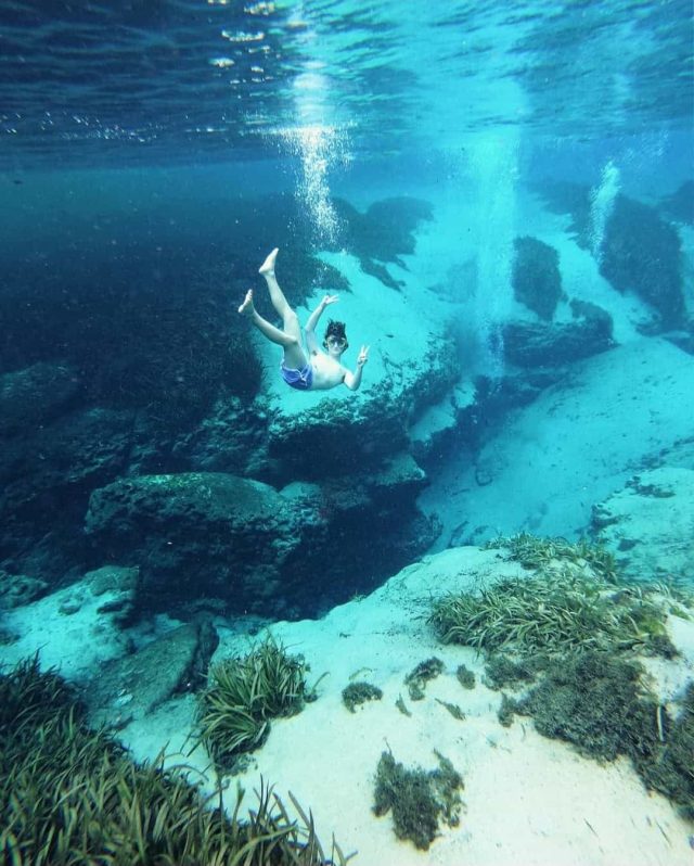 Dean Keeley submerged in clear blue water, looking at the camera and holding up peace signs with his fingers.